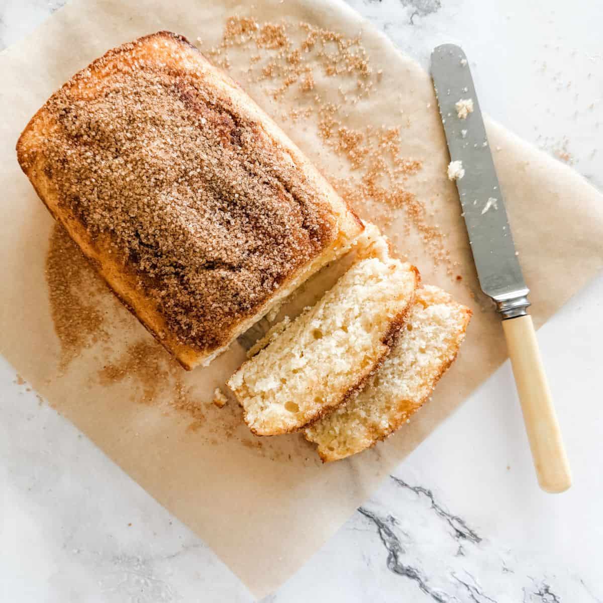 Cinnamon tea cake with two slices out on a sheet of baking paper on a white bench with a bone butter knife.