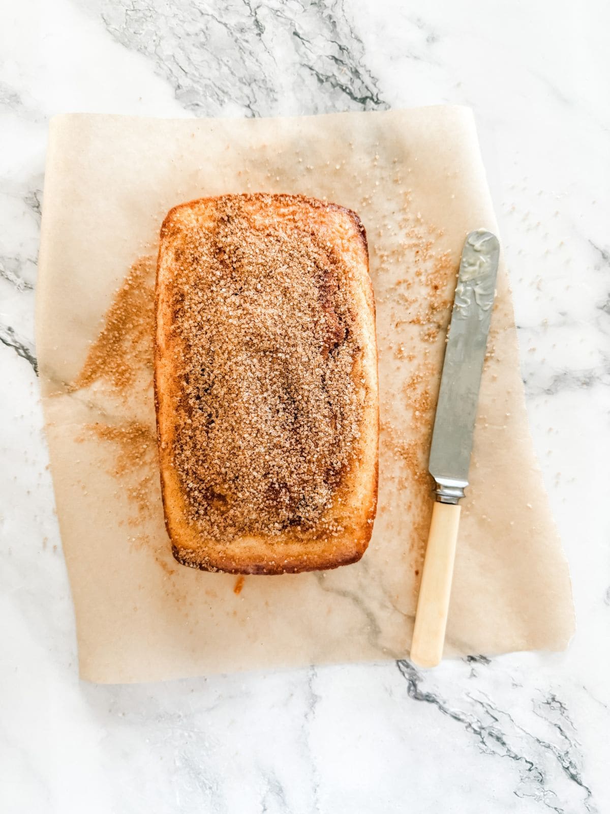 Cinnamon tea cake on sheet of baking paper with bone butter knife on white marble bench.