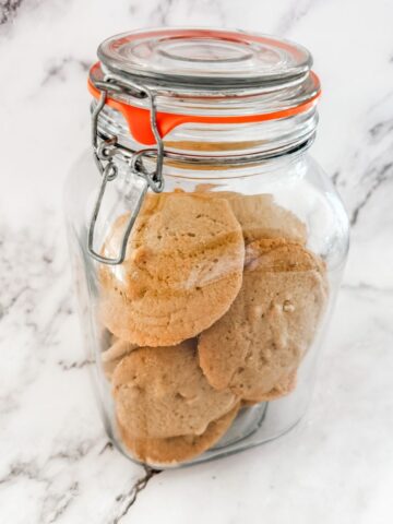 Brown sugar walnut cookies in a glass cookie jar on a white marble bench top.