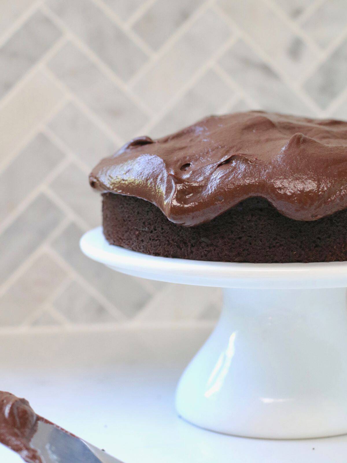 Chocolate fudge icing on top of a chocolate cake on a white stand