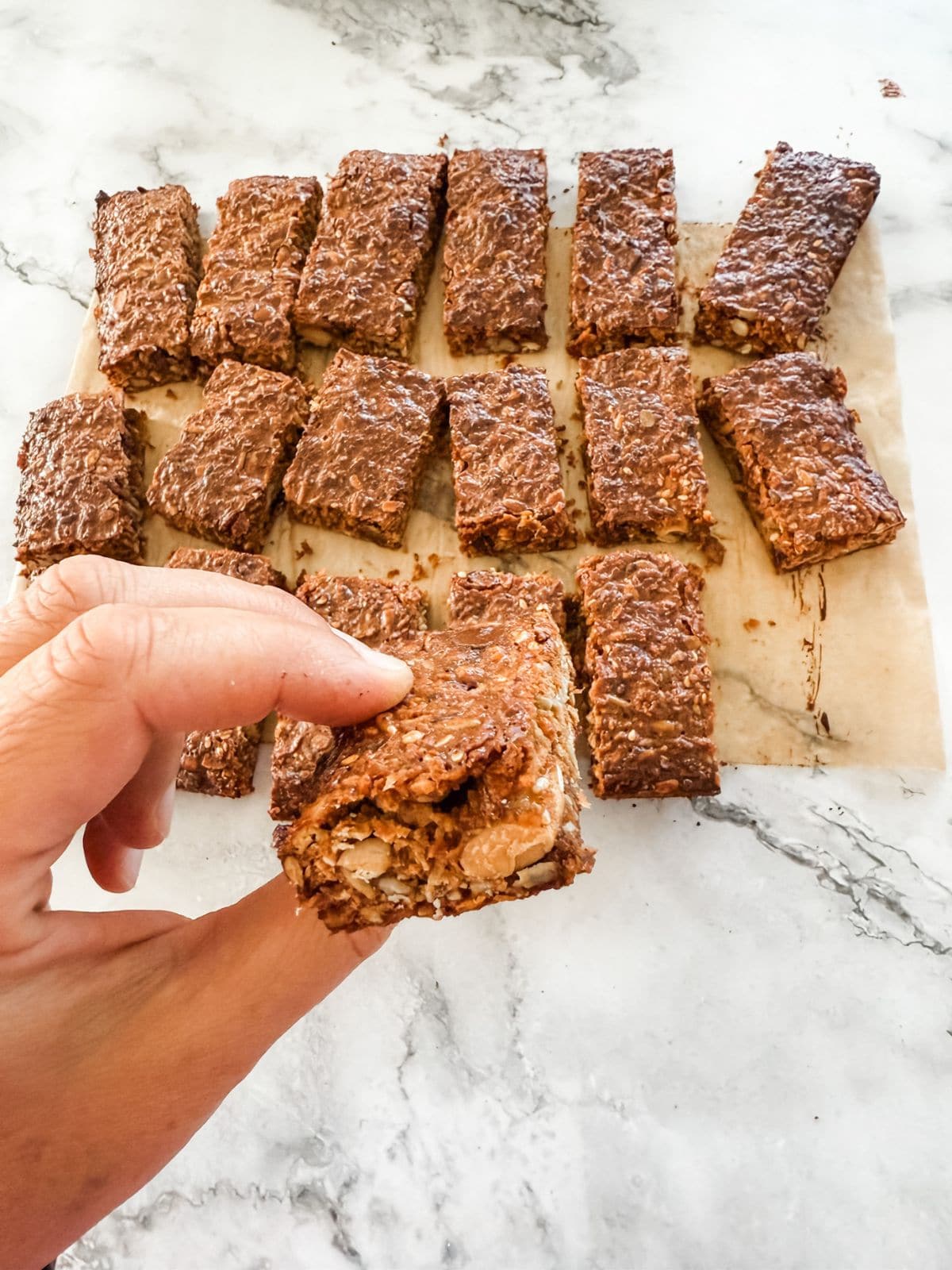 Hand holding homemade muesli bar over tray of muesli bars. 