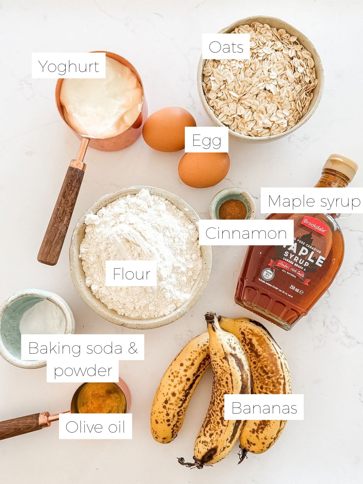 Bowl of flour, 2 bananas, 2 eggs, bottle of maple syrup, cinnamon powder, bowl of oats and baking powder to make healthy Banana Oat Muffins.