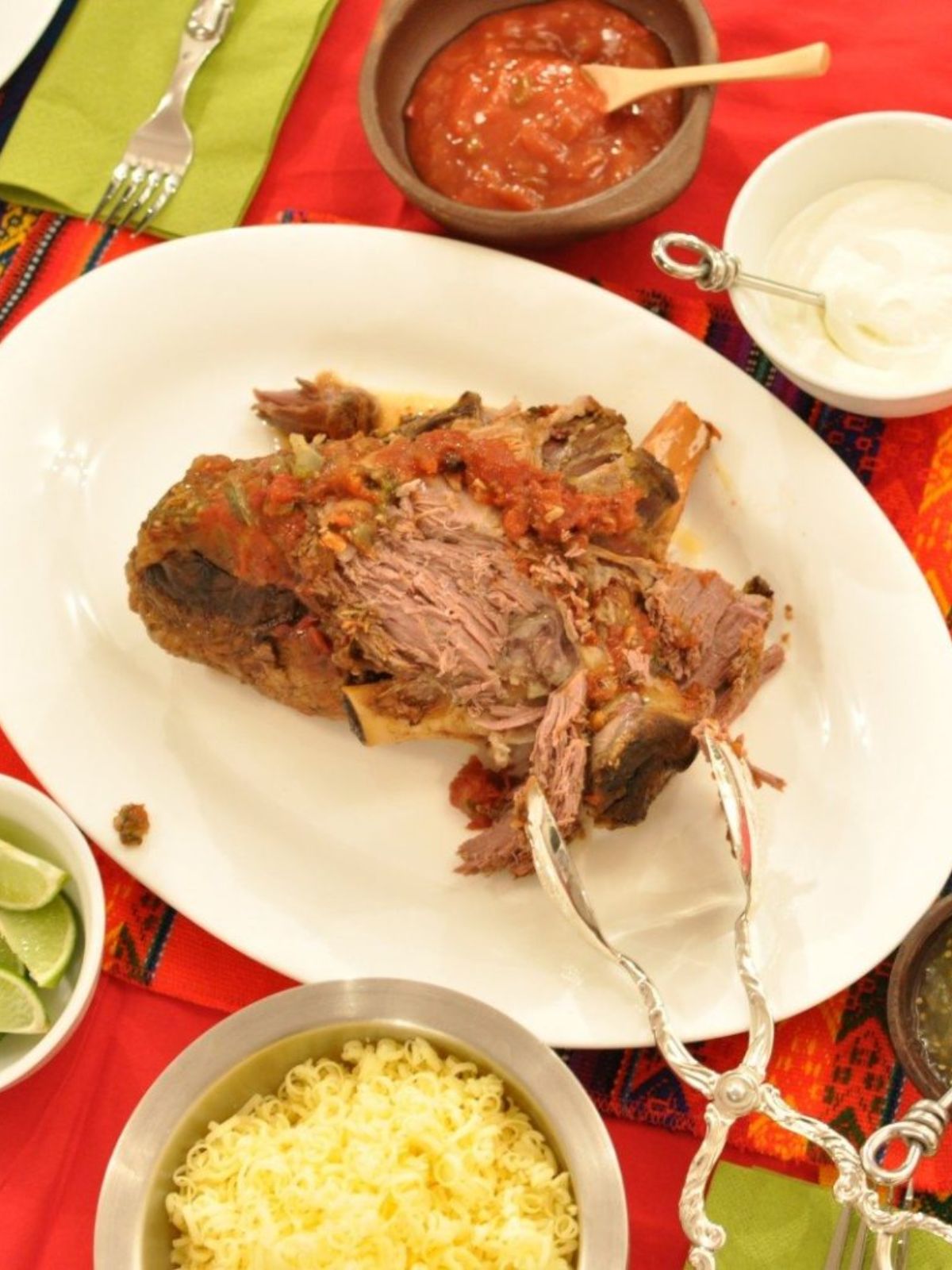Slow cooked leg of lamb partly shredded on white plate on red table cloth with bowl of cheese next to it. 