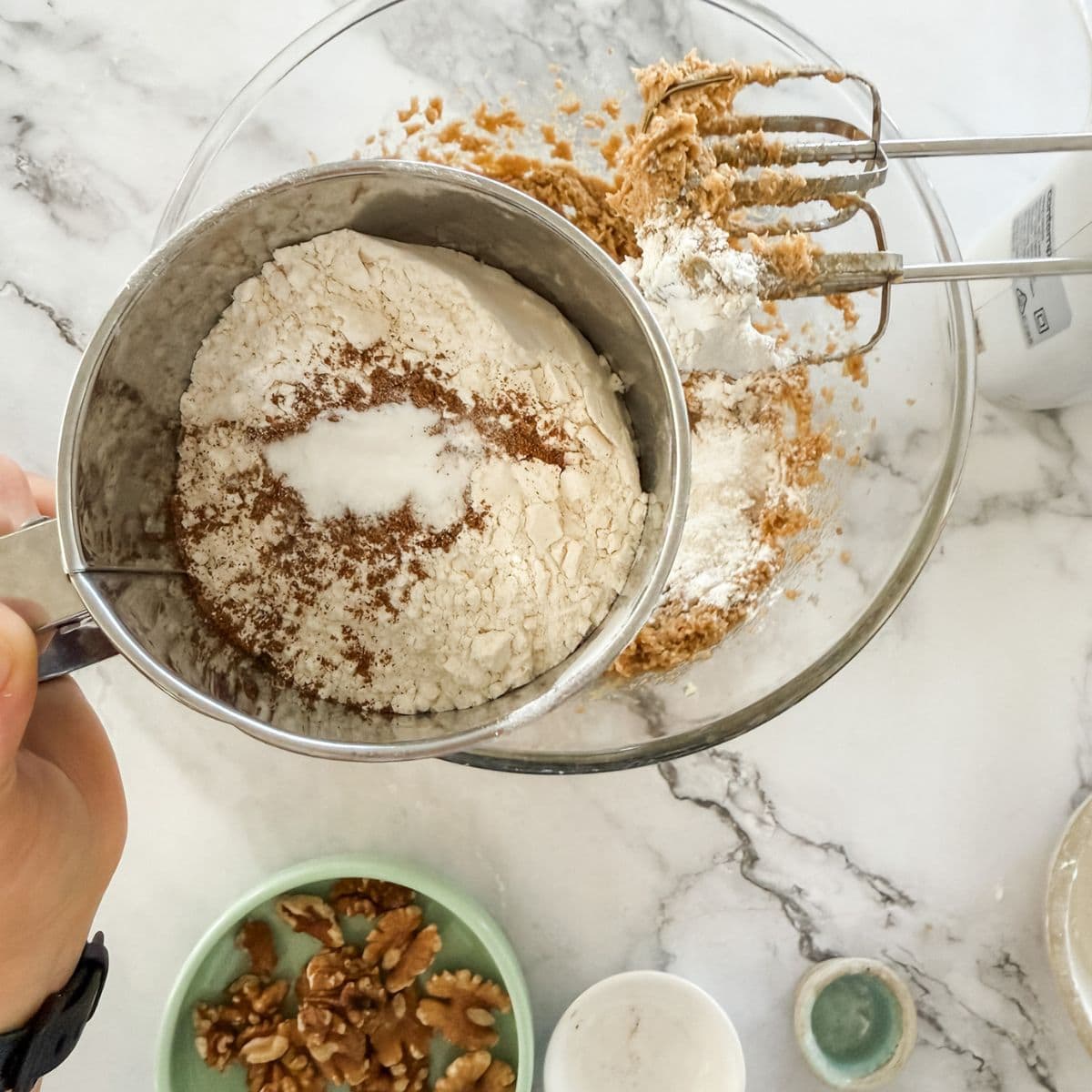 A handheld sifter sifting flour, mixed spice and baking soda into a glass bowl.
