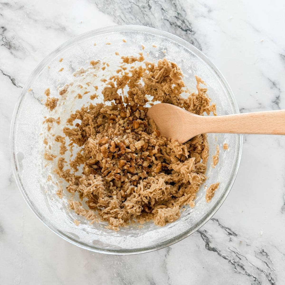 Brown cookie dough topped with chopped walnuts in a glass bowl topped with a wooden spoon on a marble bench top.