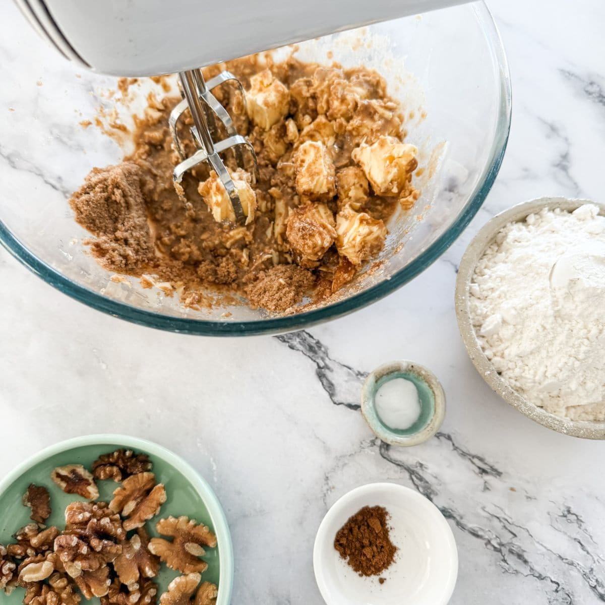 Butter, brown sugar in a glass bowl being beaten with a hand held electric mixer. Also shows bowl of walnuts, flour and mixed spice.