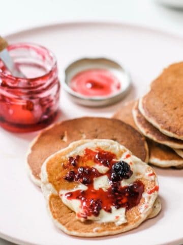 Mulberry jam and butter spread on pancakes with a small jar of jam in the background and tiny knife. All on pale pink plate