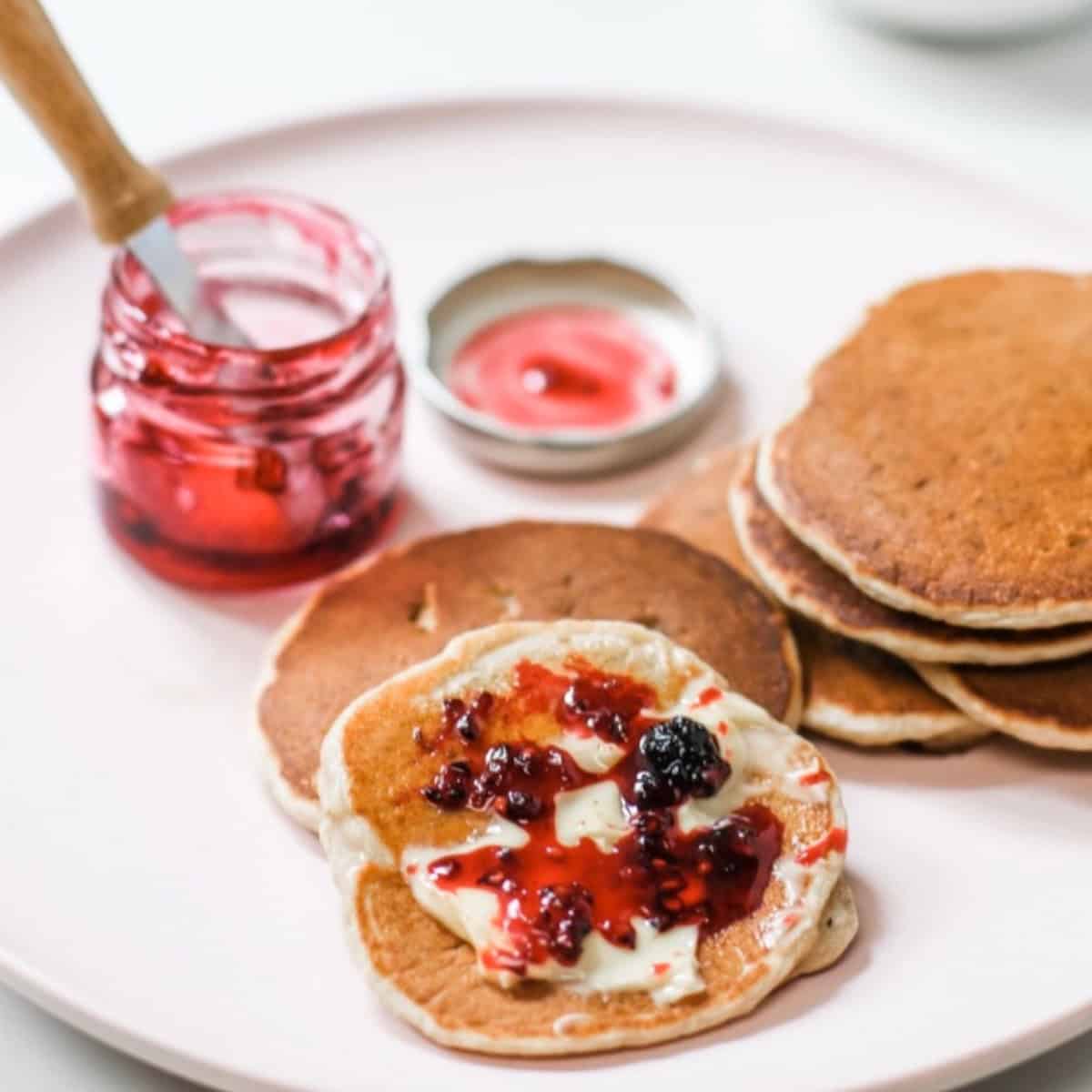Mulberry jam and butter spread on pancakes with a small jar of jam in the background and tiny knife. All on pale pink plate.