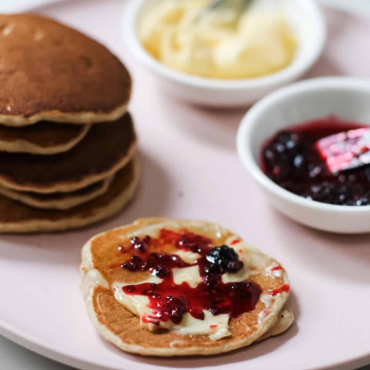 Mulberry jam and butter spread on pancakes with a small jar of jam in the background and tiny knife. All on pale pink plate