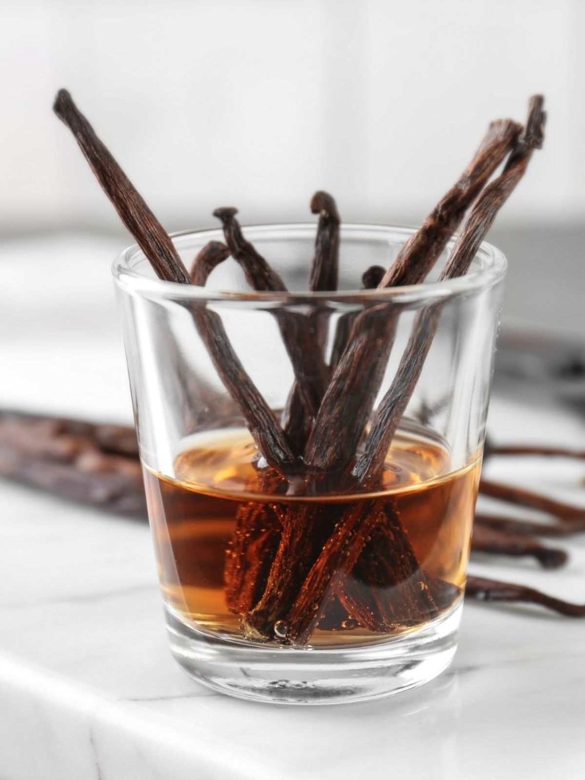 Vanilla beans in a glass jar of vanilla extract with vanilla beans in the background on a white marble surface. 