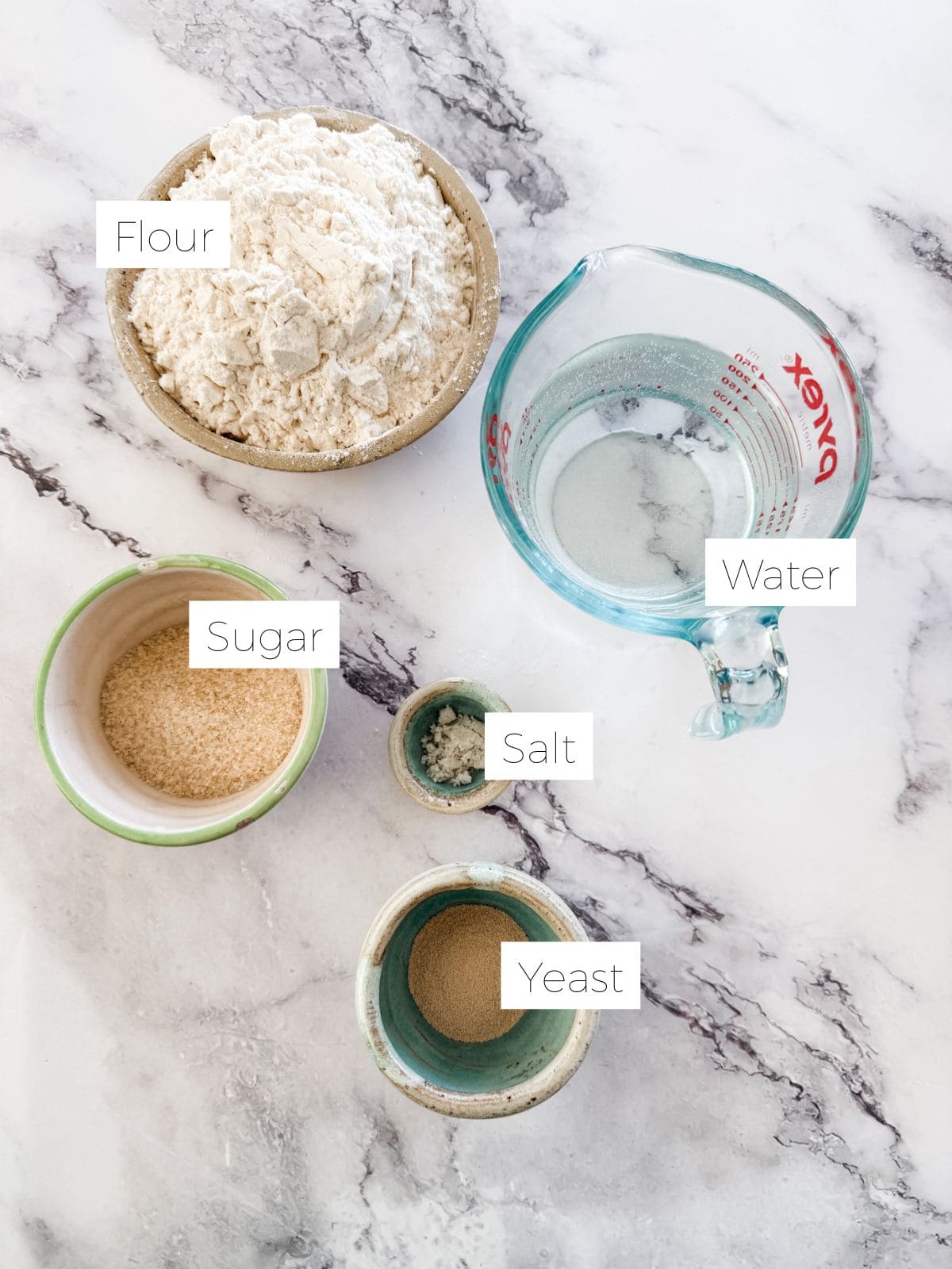 Bowl of flour, jug of water, bowl of sugar, salt & yeast on white marble bench top. 