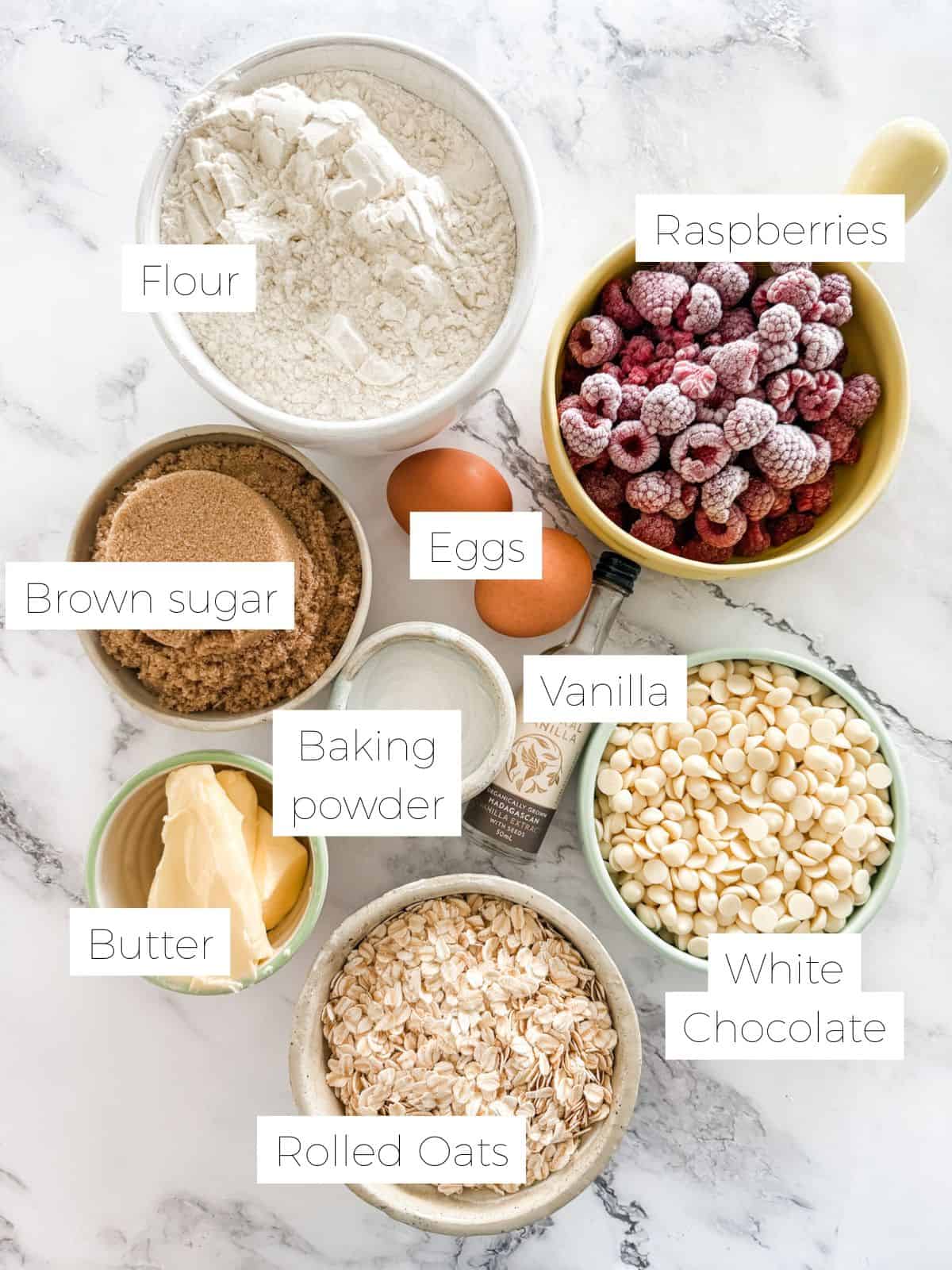 Ingredients in Healthy raspberry and white chocolate muffins - bowl of flour, raspberries, sugar, white chocolate, 2 eggs, rolled oats, butter