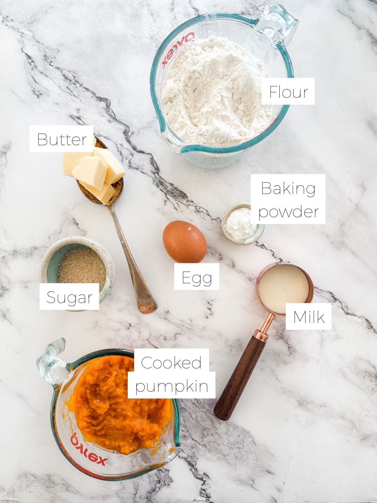 Ingredients in Classic Pumpkin Scones - flour in jug, butter on spoon, egg, milk, pumpkin puree, sugar in bowl on white bench. 