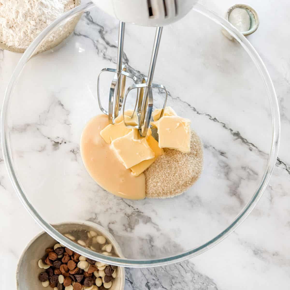 Glass bowl with butter, condensed milk, sugar & vanilla with handheld beater on white marble bench.