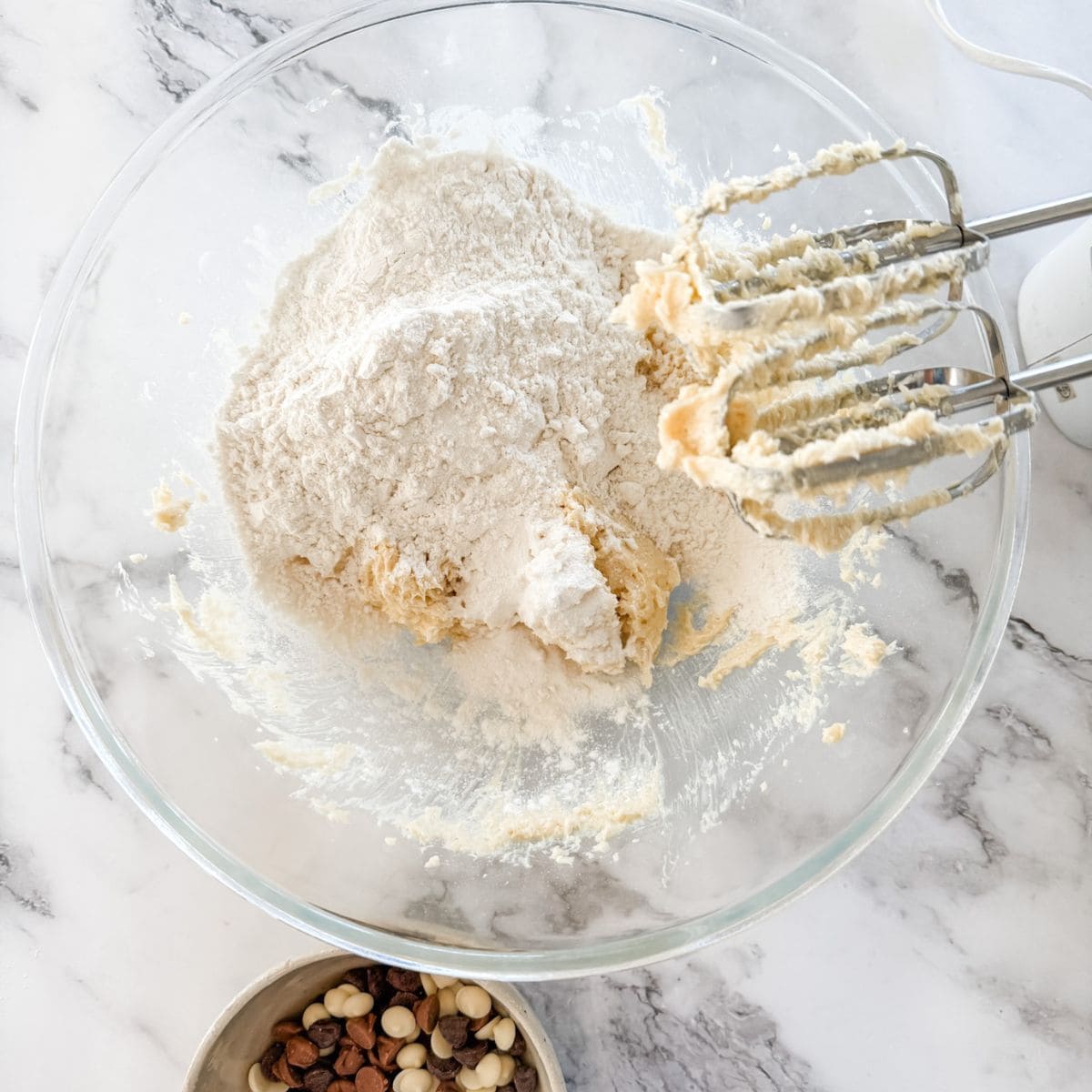 Flour on top of biscuit batter with handheld beaters in glass mixing bowl.