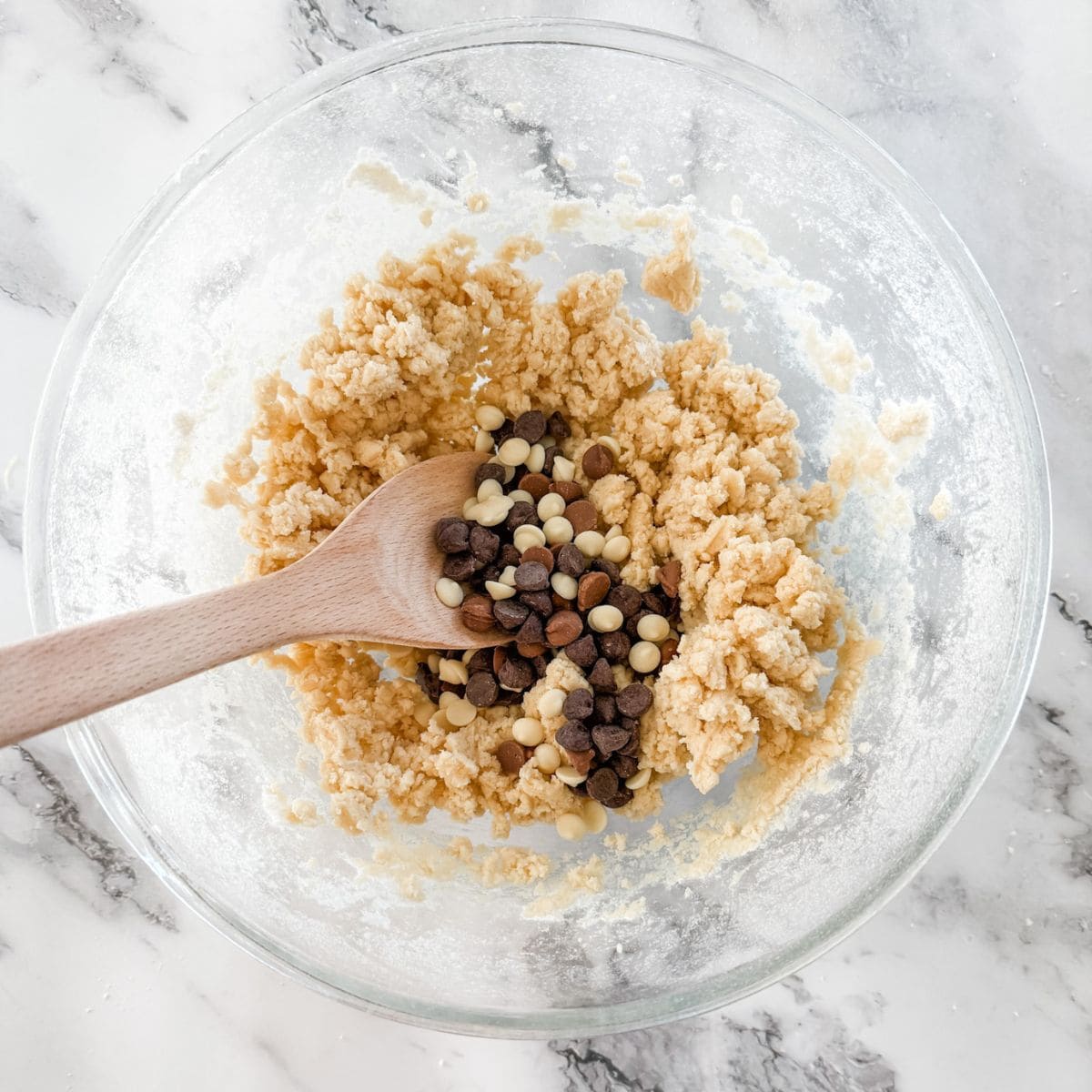 Chocolate chips on top of cookie batter with wooden spoon in glass mixing bowl on white marble bench top.