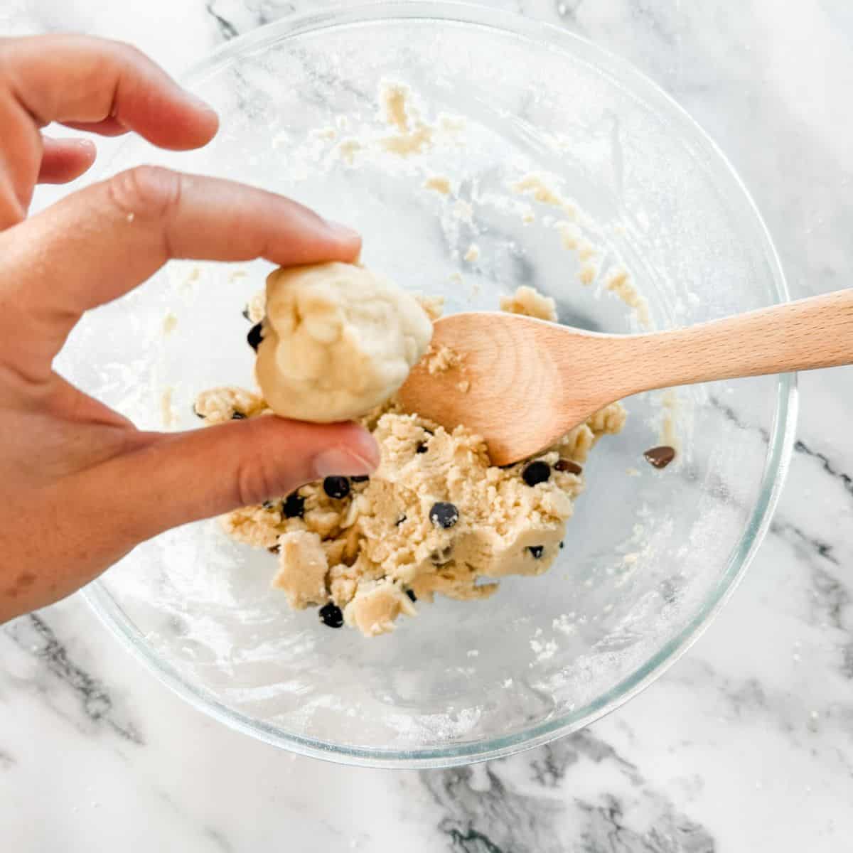 Hand holding ball of chocolate chip cookie dough over glass mixing bowl of dough on white marble bench.