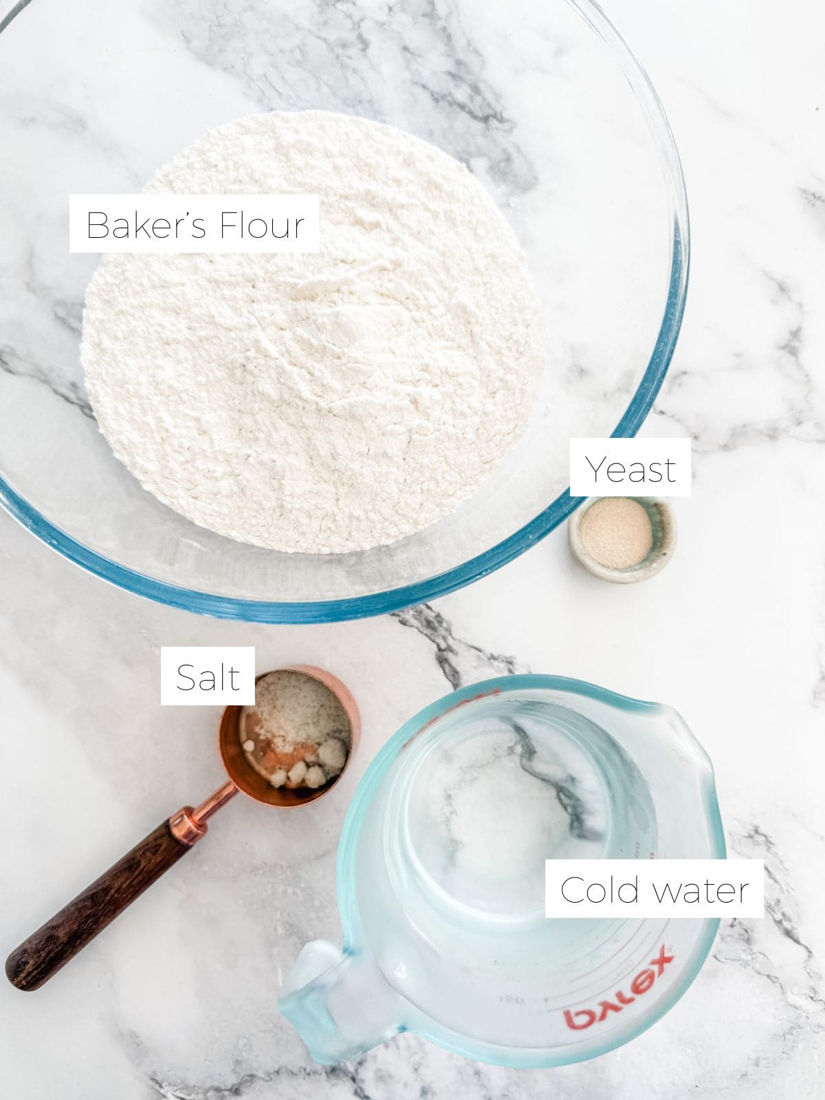 Large glass bowl of flour, measuring cup of water, bowl of salt, measuring cup of yeast.