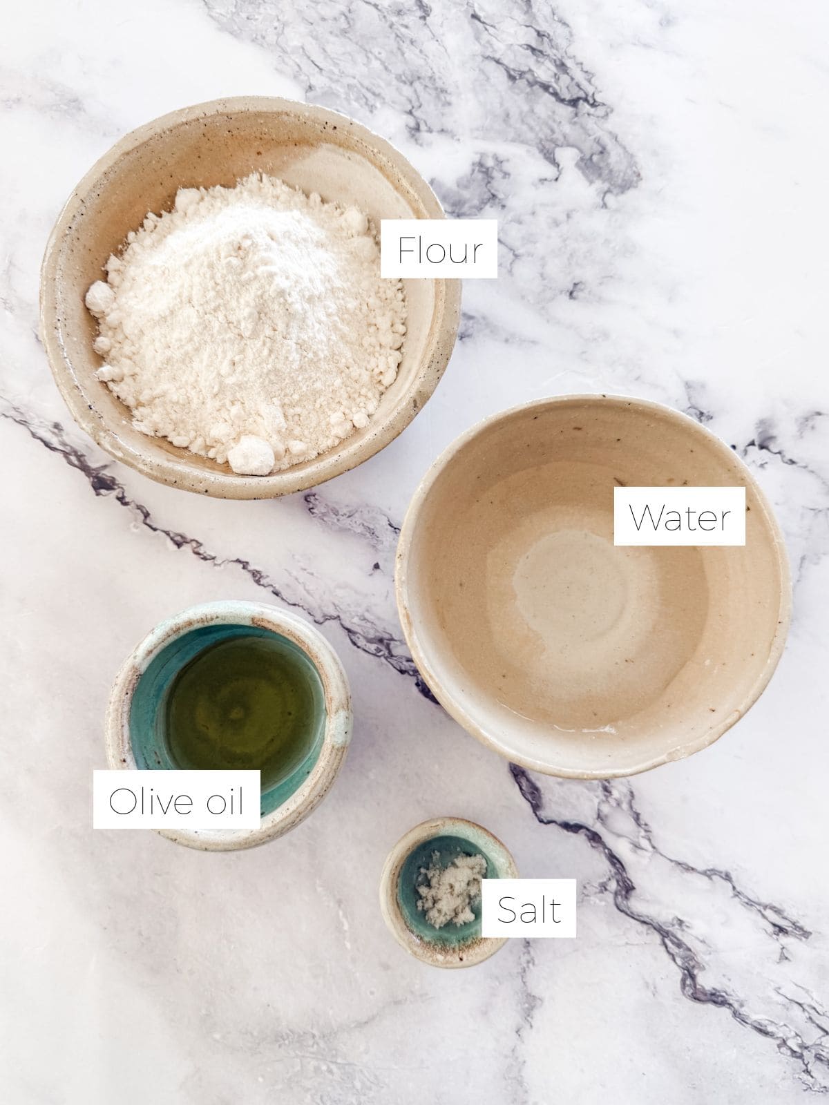 Bowl of flour, water, olive oil, salt on white marble bench top.