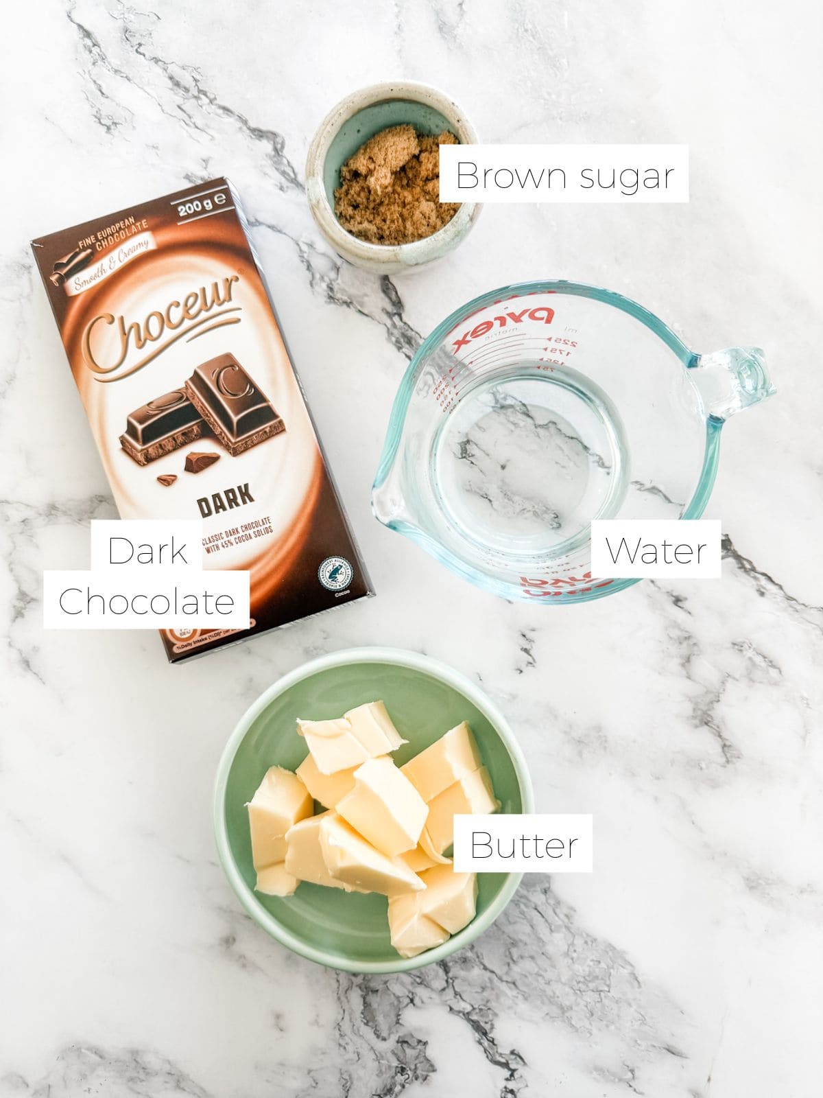Ingredients in Best chocolate icing - bowl of brown sugar & bowl of butter, jug of water and block of dark chocolate on white bench top.