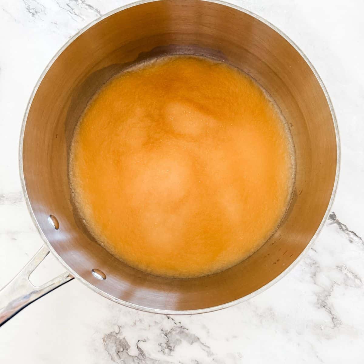 Stainless steel pot with juice, bloomed gelatine and honey on a white marble bench top - making jelly lollies.