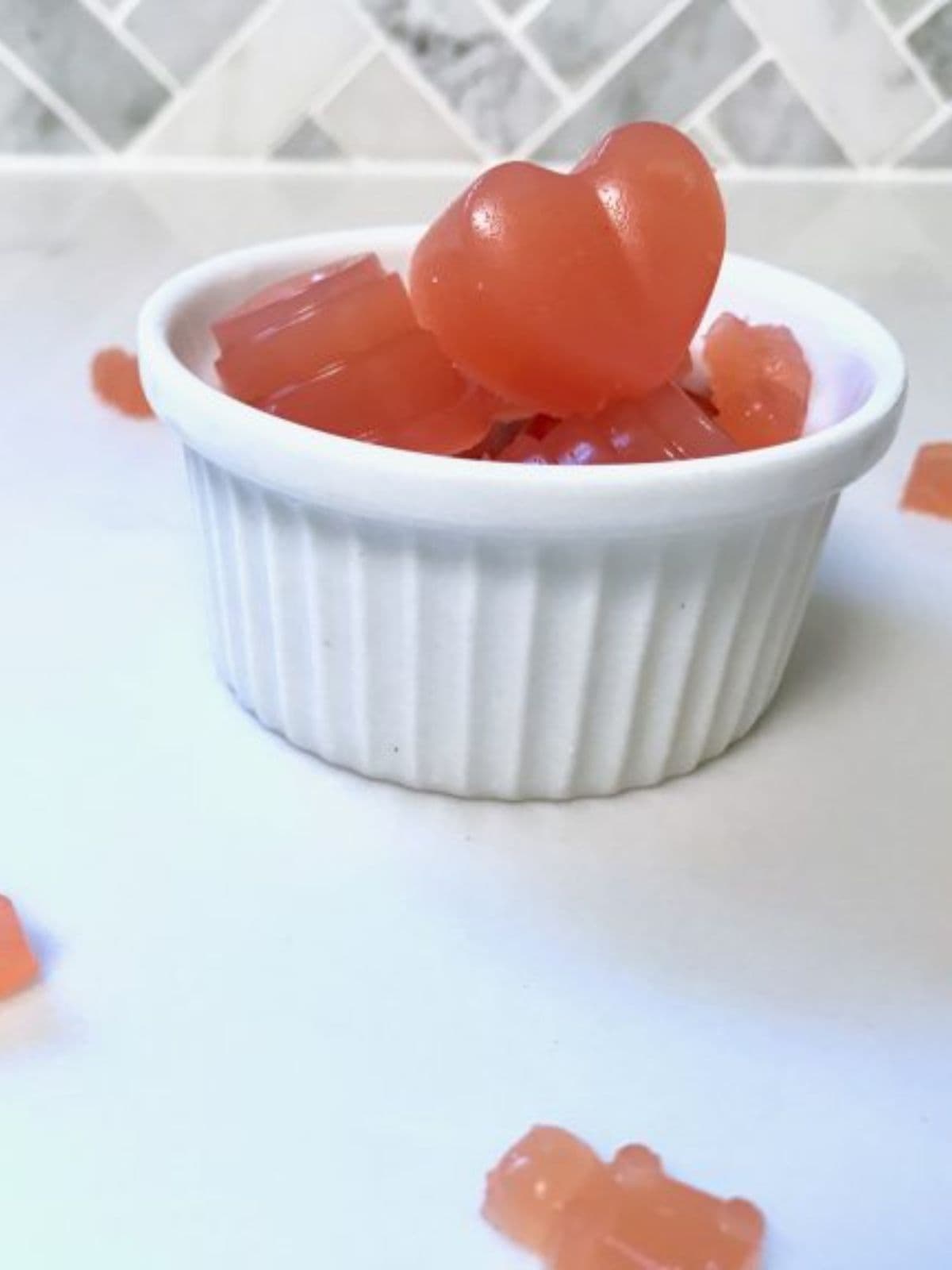 Heart shaped jelly lollies in a white ramekin on a white benchtop with homemade gummy bears spread around.