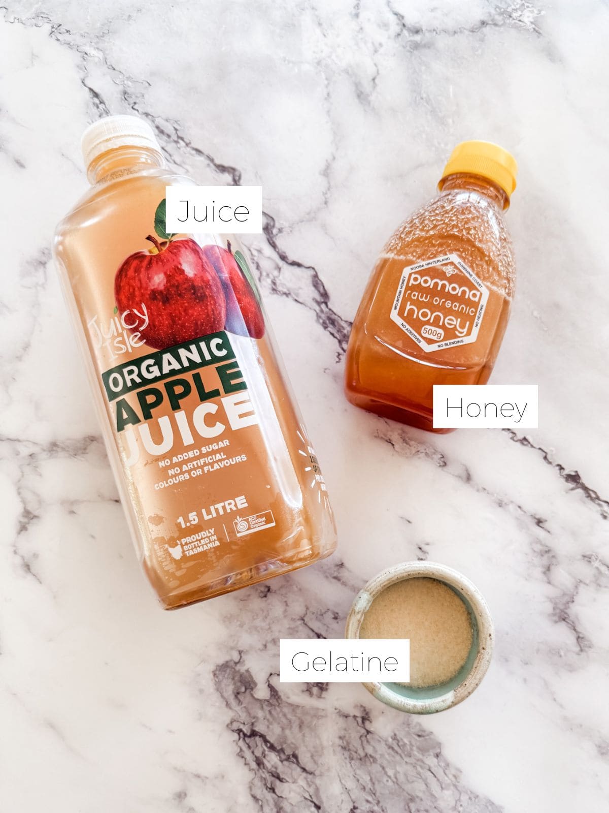 Ingredients for making jelly lollies. Bottle of apple juice, squeeze bottle of honey and small bowl of gelatine powder on white marble bench. 