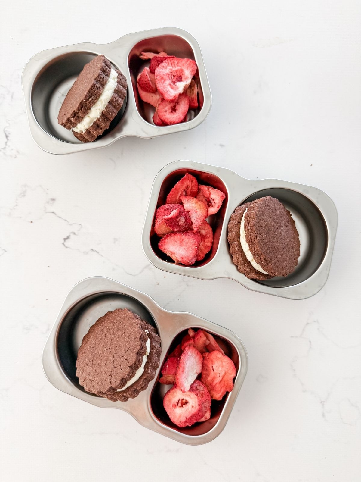 Chocolate biscuits sandwiched with white icing in a lunchbox with freeze dried strawberries. 